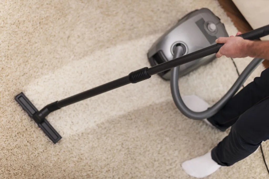 Person vacuuming a beige carpet with a gray vacuum cleaner. The scene shows a clean, orderly home environment focused on tidiness and maintenance.