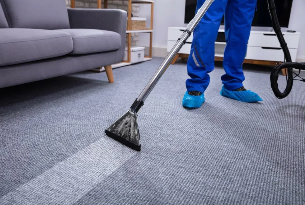 Professional carpet cleaning in a modern home worker in blue uniform using a carpet extractor to deep clean gray carpet with visible results.